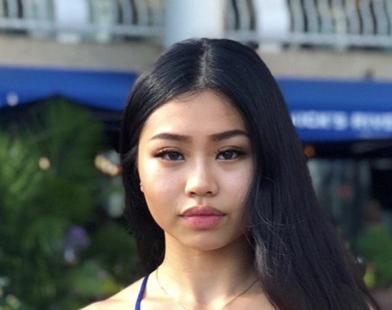 Image of a young woman with long black hair, wearing a blue top, standing outdoors with greenery and a building with a blue awning in the background.