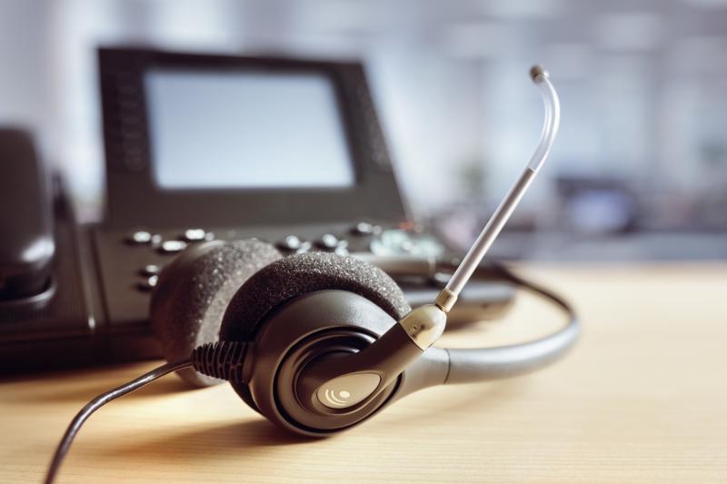 A headset with a microphone resting on a wooden desk, next to a telephone with a small screen, set against a blurred office background.