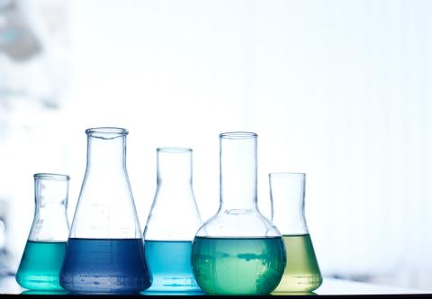 A row of laboratory flasks containing colored liquids, including blue, green, and yellow, set against a blurred background.